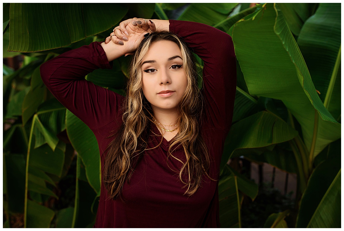 girl places hands over head in front of large green leaves during senior portraits