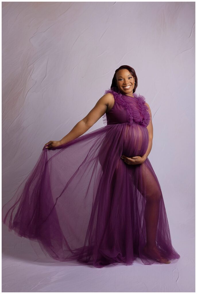 woman wears sheet purple gown as she holds her pregnant belly and smiles by Amy Yang Photography